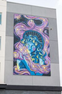 Mural on a building wall depicting an individual with flowing hair, surrounded by stars and cosmic patterns in purple, blue, and yellow tones.